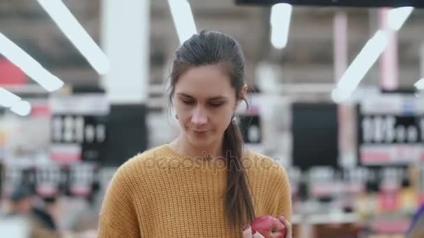 Вид спереди женщины, выбирающей яблоки в супермаркете, держащей яблоко. Молодая девушка рассматривает фрукты. 4K — стоковое видео