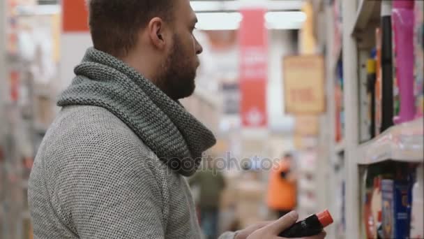 El joven que elige el artículo en los estantes de la tienda, quiere comprar productos químicos para el hogar. 4K — Vídeos de Stock