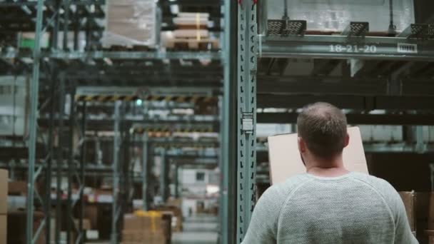 Un homme prend des boîtes des étagères, les prend et s'en va à travers des étagères dans un entrepôt. Vue de derrière. 4K — Video