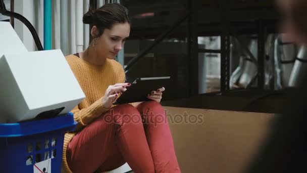 Jovem mulher sentada no armazém e usando tablet para verificar a lista de mercadorias. O homem com o carrinho vai para a frente da câmara. 4K — Vídeo de Stock