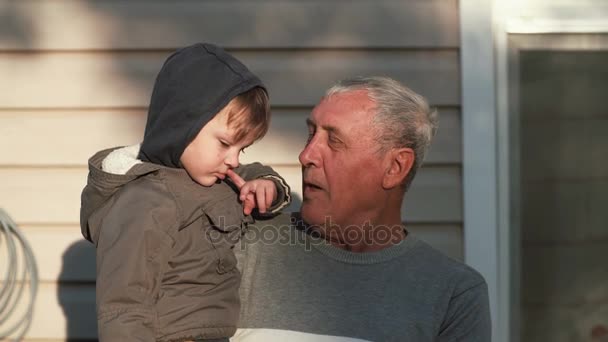 Старик держит за руку маленького мальчика. Дедушка и внук разговаривают, улыбаются, смотрят на камеру под открытым небом на солнце. 4K — стоковое видео