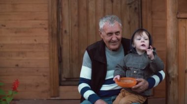 Dedesi s kucağına oturan ve turuncu kaseden meyveleri yemekten şirin küçük çocuk. Aile hafta sonu açık. 4k