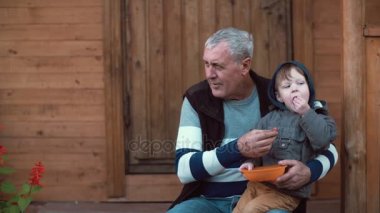 Dedesi s kucağına oturan ve turuncu kaseden meyveleri yemekten şirin küçük çocuk. Yaşlı adam besleme torunu. 4k