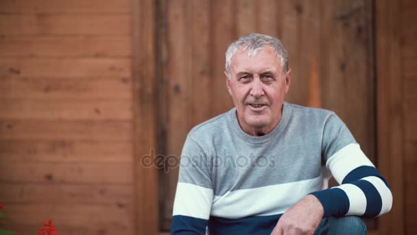 Velho sentado no alpendre da casa. O pensionista vira a cara e olha para a câmara, sorrindo, falando. 4K — Vídeo de Stock