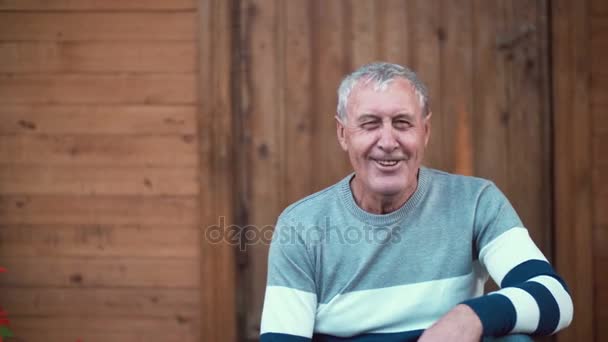 Öreg ember ül a tornácon a ház. Nyugdíjas keres, hogy a kamera, nevetve, mosolyogva, beszél. 4k — Stock videók