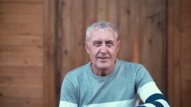 Portrait du vieil homme assis sur le porche de la maison. Pensionné regardant la caméra, souriant et fumant. 4K — Video