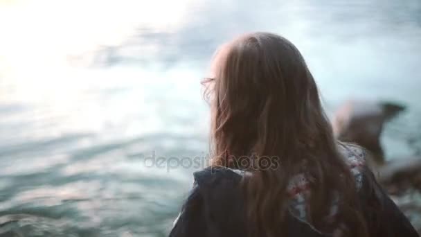 Menina bonito em pé na costa perto da água e joga uma pedra na água . — Vídeo de Stock