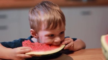 Yakın çekim görünümünü mutfak masada oturan sevimli küçük çocuk. Bir parça tutan ve bir karpuz yeme çocuk.