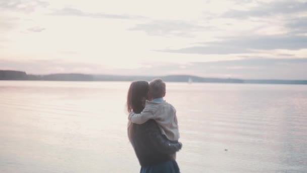 Young brunette woman holding little boy on the coast of the beach. Mother kiss her son, pointing with finger at horizon. — Stock Video