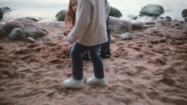 Kıyıda oturan ve alır oynayan sevimli küçük kız el kum ve yere dökülen. Kardeş kardeş için gidiş. — Stok video