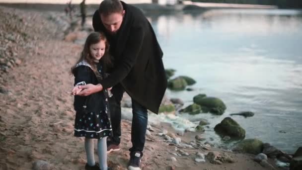 Батько і дочка на березі, маленька дівчинка кидає камінь у воду. Чоловік показує дівчині, як грати пропускає камінь . — стокове відео