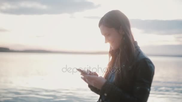 Молода красива жінка стоїть на узбережжі і використовує смартфон. Дівчина на пляжі зворушливий екран і посмішка . — стокове відео