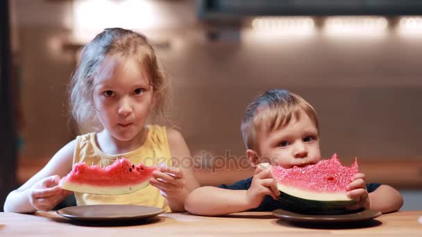 Bruder und Schwester sitzen am Tisch in der Küche. Junge und Mädchen essen frische Wassermelone und schauen in die Kamera. — Stockvideo