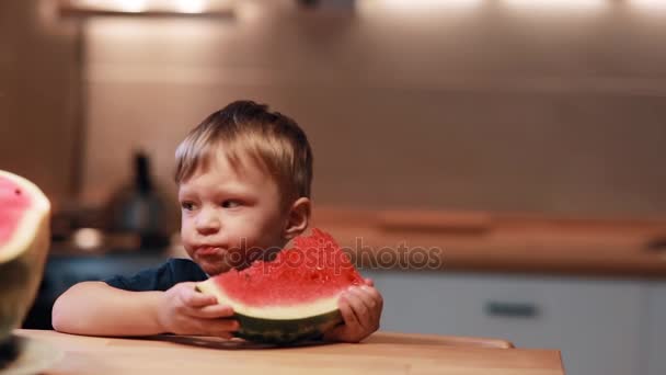 Porträt des netten kleinen Jungen, der am Tisch in der Küche sitzt. ein Stück halten und eine Wassermelone essen. — Stockvideo