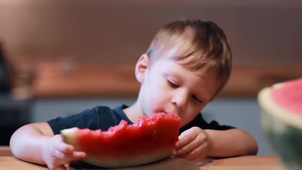 Vista de cerca del lindo niño sentado en la mesa de la cocina. Macho sosteniendo una pieza y comiendo una sandía . — Vídeos de Stock