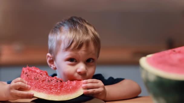 Close-up beeld van aantrekkelijke jongetje knabbelen de watermeloen, zitten aan de tafel. Mannelijke open mond en beet een stuk. — Stockvideo