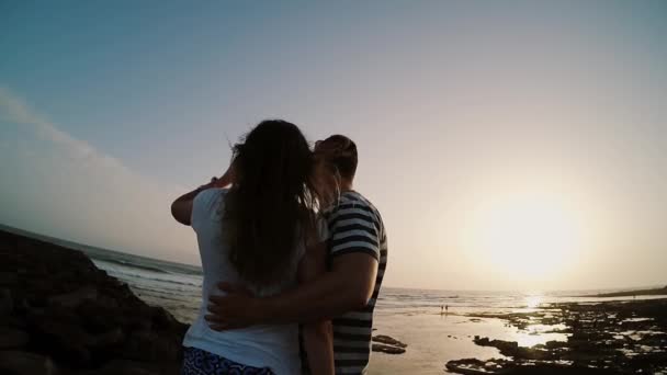 Hombre y mujer de pie en la orilla junto al mar, abrazándose y mirando al atardecer. Pareja en la playa de la tarde. Mociones lentas . — Vídeo de stock