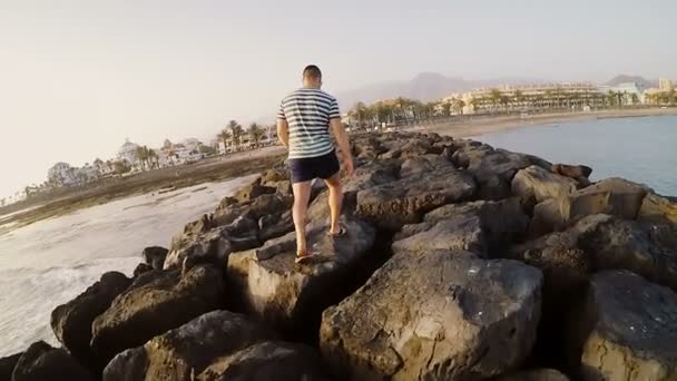 Vista trasera del joven caminando a través de las rocas en la playa. Hombre pasando tiempo fuera cerca del mar en un día soleado . — Vídeos de Stock