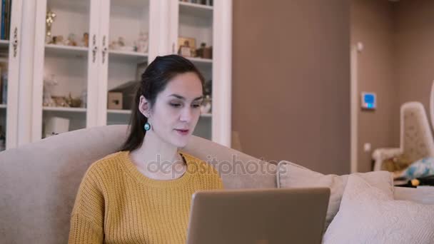 Jovem mulher sentada no sofá e segurando laptop. Feminino tem uma videoconferência, falando na Internet . — Vídeo de Stock