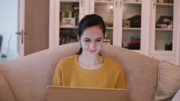 Junge Frau mit Laptop, während sie im Wohnzimmer sitzt. Frau sitzt auf Sofa und arbeitet am Computer und lächelt. — Stockvideo