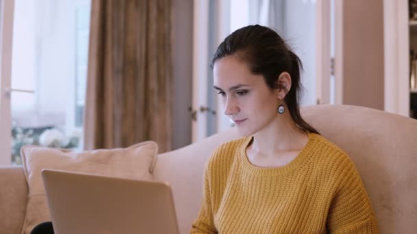 Mujer joven sentada en el sofá en la sala de estar y utilizando el ordenador portátil. Mujer navegando por Internet y sonriendo . — Vídeos de Stock