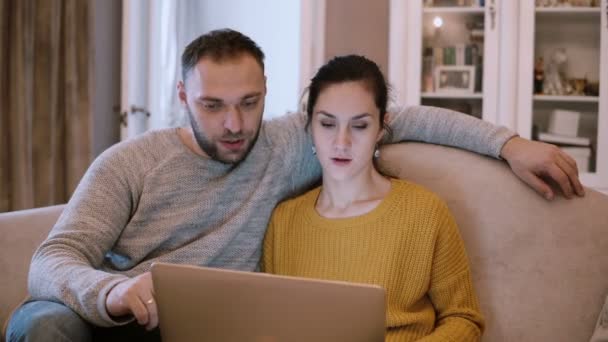 Νεαρό ζευγάρι κάθεται στον καναπέ και κρατώντας τον υπολογιστή. Άνδρας και γυναίκα που χρησιμοποιούν φορητό υπολογιστή ενώ κάθεται στο σαλόνι. — Αρχείο Βίντεο