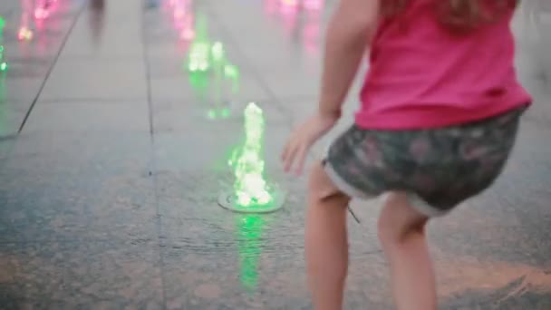 Schattig klein meisje speelt met gekleurde water jets bij de fontein in warme zomerdag. — Stockvideo