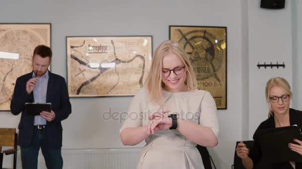 Mujer rubia joven pasando por la oficina moderna y mirando el reloj inteligente. La chica usa nueva tecnología. Movimiento lento — Vídeos de Stock