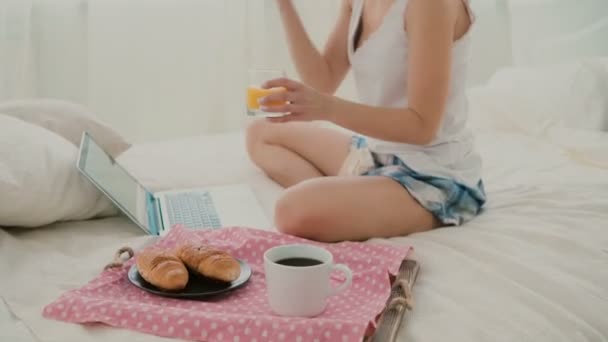 Junge attraktive Frau trinkt Saft auf dem Bett. Brünettes Mädchen surft beim Frühstück mit Laptop im Internet. — Stockvideo