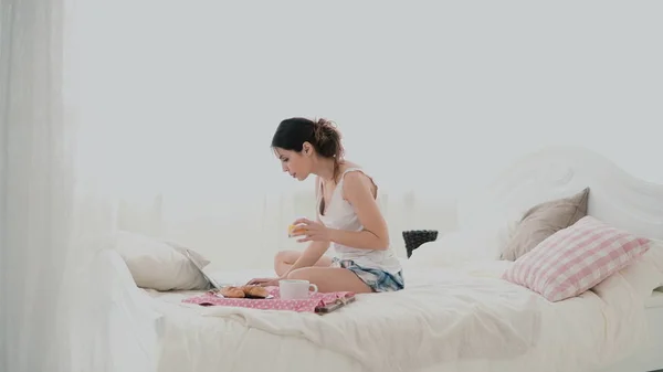 Fiatal barna nő húzódik egy ágyban. Gyönyörű lány használ laptop, reggeli közben. Ivás lé és a tánc. — Stock Fotó