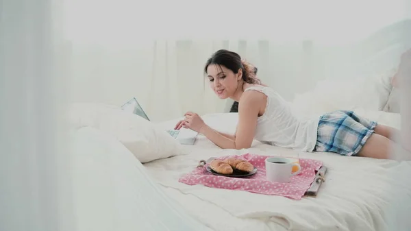 Fiatal nő használ laptop ágyon fehér otthon reggeli közben. Boldog lány enni, croissant és tallózások az interneten. — Stock Fotó