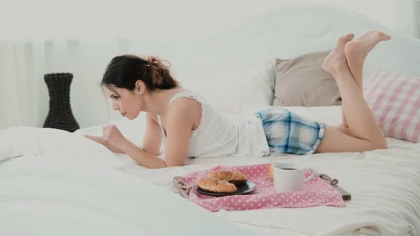 Schönes Mädchen, das morgens auf dem Bett liegt und plaudert und dafür Laptop benutzt. junge Frau isst Croissant und tippt. — Stockfoto