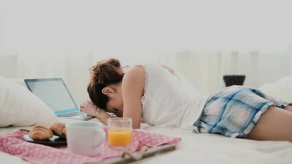 Brunette girl lying on bed in morning and chatting, using laptop for that. Young woman typing on keyboard and laughing. — Stock Photo, Image