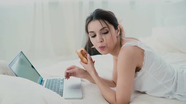 Beautiful girl lying on the bed in morning, using laptop. Young woman eating croissant, typing, browsing the Internet. — Stock Photo, Image