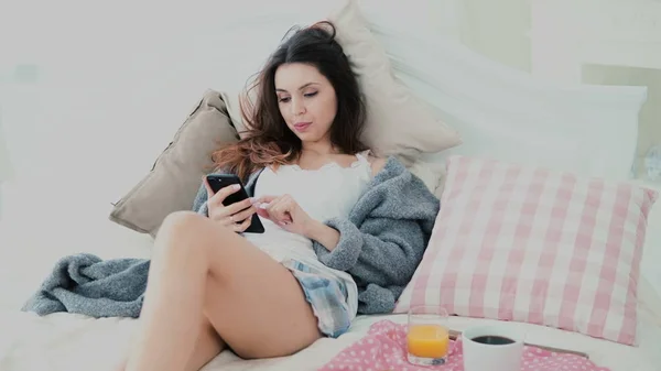 Junge attraktive Frau trinkt Saft auf dem Bett. Brünettes Mädchen surft während des Frühstücks mit Smartphone im Internet — Stockfoto