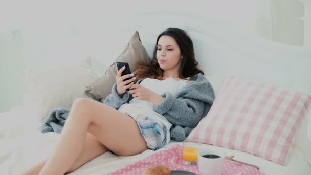 Young woman using smarphone during breakfast lying on bed at home. Attractive girl surfs the Internet on mobile phone. — Stock Video