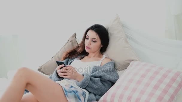 Mooie brunette vrouw gebruik van smartphone, touchscreen vertoning op lichte kamer. Meisje ligt op bed en op het Internet surft. — Stockvideo