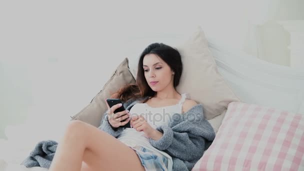 Hermosa mujer usando teléfono inteligente, pantalla táctil en la sala de luz. Chica se encuentra en la cama, navega por Internet y sonríe . — Vídeos de Stock