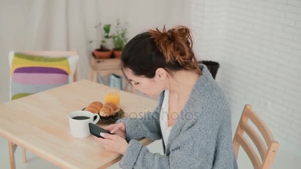 パジャマを着て、インター ネット サーフィン、キッチンに座っているかわいい女性。ブルネットの少女は、朝食時にスマート フォンを使用してください。. — ストック動画