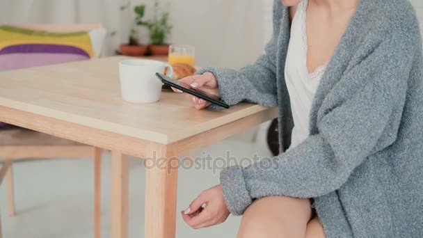 Close-up view of woman having breakfast in kitchen and browsing Internet, using smartphone. Girl chats, drinks coffee. — Stock Video