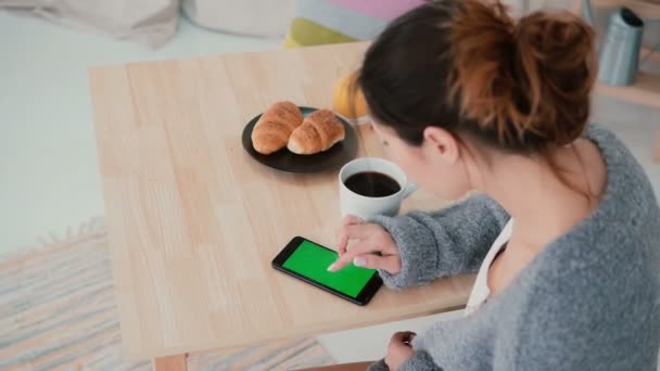 Mujer joven sentada a la mesa y tomando café en la cocina. Chica morena utiliza smartphone, pantalla verde . — Vídeos de Stock