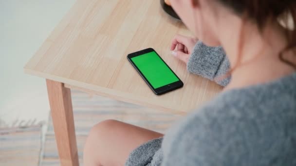 Zadní pohled na ženu sedící u stolu v kuchyni doma. Tmavovláska používá smartphone, zelená obrazovka. — Stock video