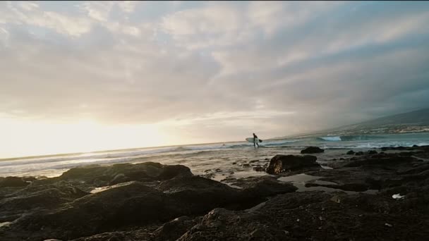 Blick auf den Sonnenuntergang am Strand. Surfer, der ein Surfbrett hält und ins Wasser kommt. wunderschöne Meereslandschaft. — Stockvideo