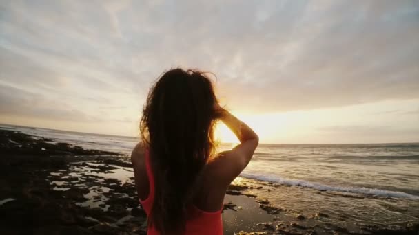 Молодая женщина на пляже смотрит на красивый закат. Женщина наблюдает за горизонтом, ветер дует волосами. Наслаждаясь морем . — стоковое видео