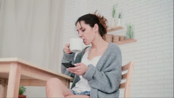 Hermosa mujer sentada en la cocina, navegando por Internet y desayunando. Chica utiliza el teléfono inteligente y bebe café . — Vídeo de stock