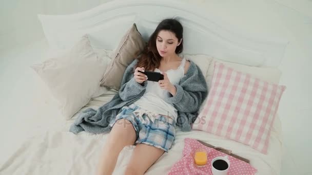 Junge schöne Frau auf dem Bett liegend und mit Smartphone. Brünettes Mädchen surft morgens beim Frühstück im Internet. — Stockvideo