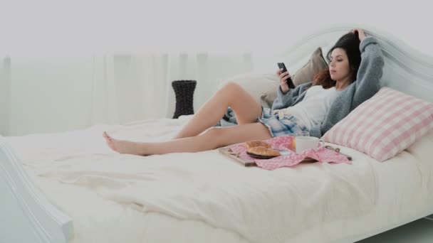 Hermosa chica acostada en la cama por la mañana y usando un teléfono inteligente. Mujer joven escribiendo, navegando por Internet y bostezando . — Vídeo de stock