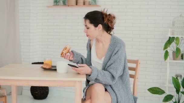 Mujer joven desayunando en la cocina y navegando por Internet, usando smartphone. Chica come el croissant, bebe café . — Vídeo de stock