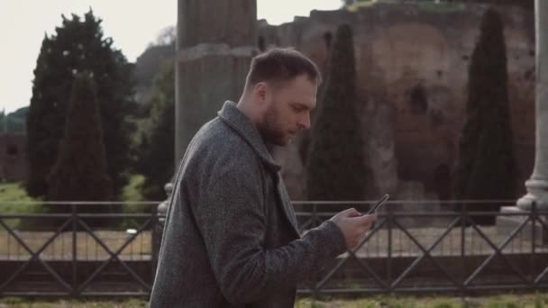 Stilig ung promenader på ruinerna av Rom, Italien. Man tittar runt romerska forumet, med hjälp av smartphone. Slow motion. — Stockvideo