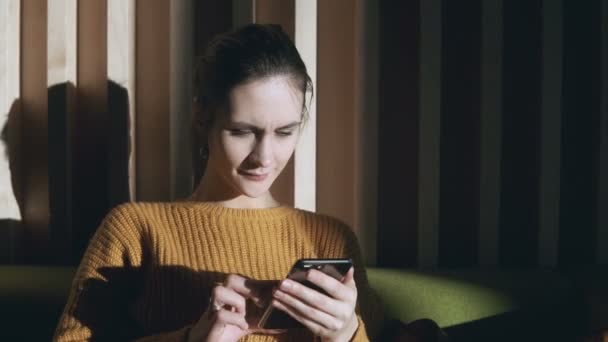 Mujer joven sentada en el sofá en la sala de estar y usando un teléfono inteligente. Mujer navegando por Internet y sonriendo . — Vídeos de Stock